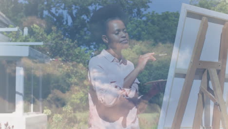 animación de nubes sobre mujer afroamericana pintando en caballete en el jardín.