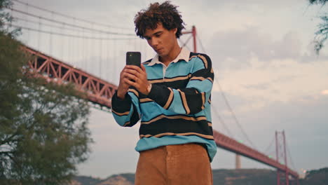 Lächelnder-Typ-Macht-Selfie-An-Der-Brücke.-Stilvoller-Hipster-Nutzt-Handy-Vertikal