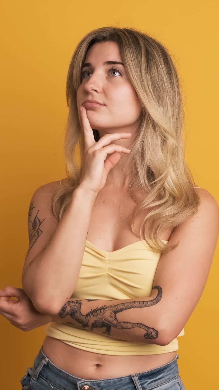 Thoughtful young woman standing with hand at chin on yellow background