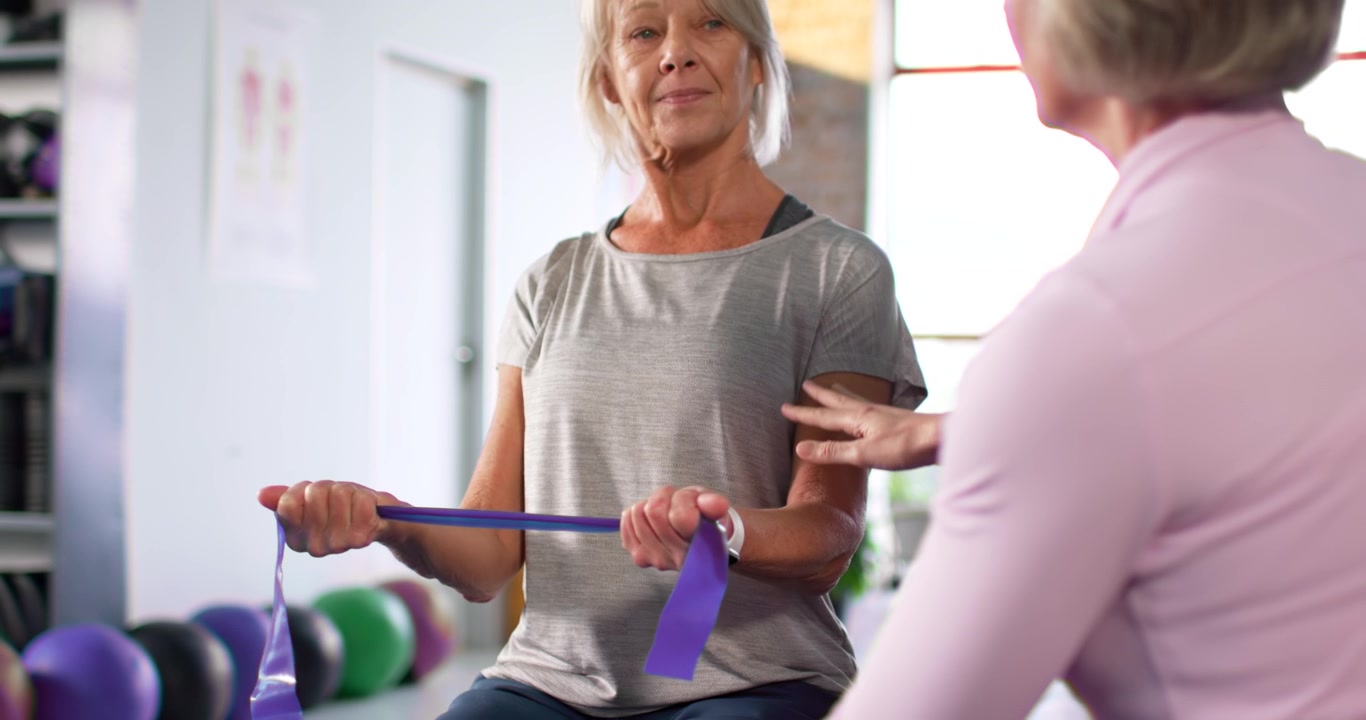 Female trainer placing hand on shoulder, knee, guiding client pulling band on ball for rehab