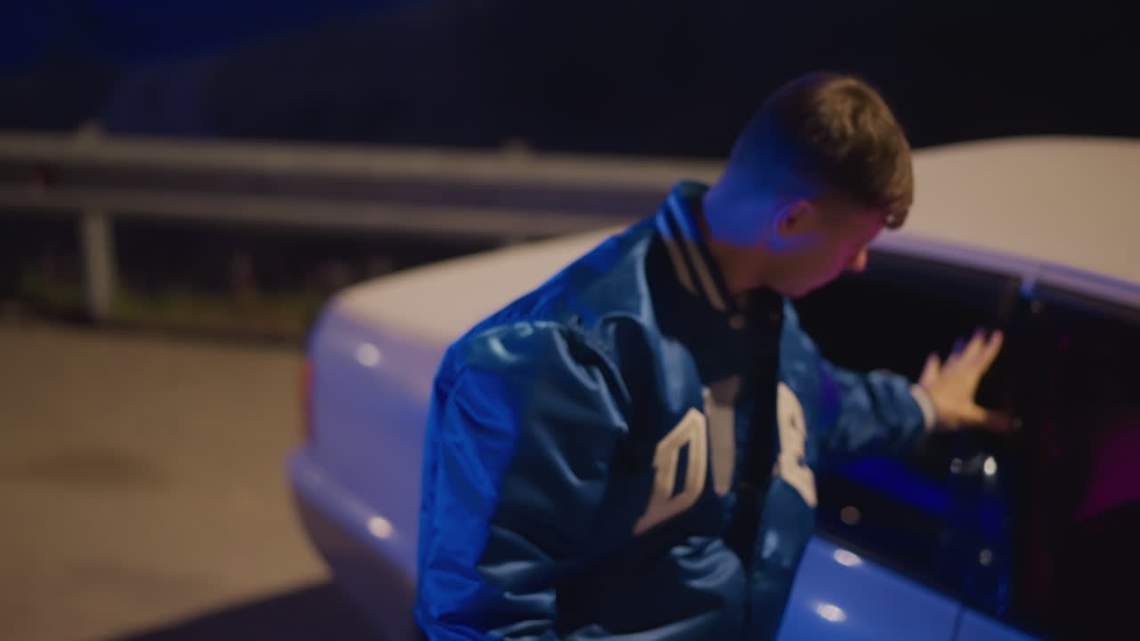 Hvid mandlig chauffør ved vejkanten, neonfarver fremhæver rat og interiør, stiger ud for at stå ved autoværnet og betragte den mørke motorvej. Universitetsjakken blafrer, udtrykket er splittet mellem at tage afsted