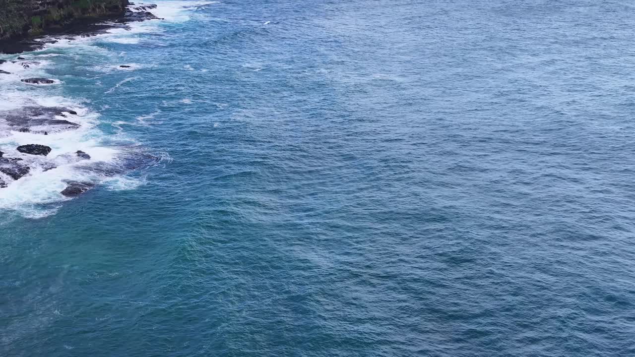 Drone camera glides above rocky shoreline, capturing blue ocean waves and rugged coastal cliffs