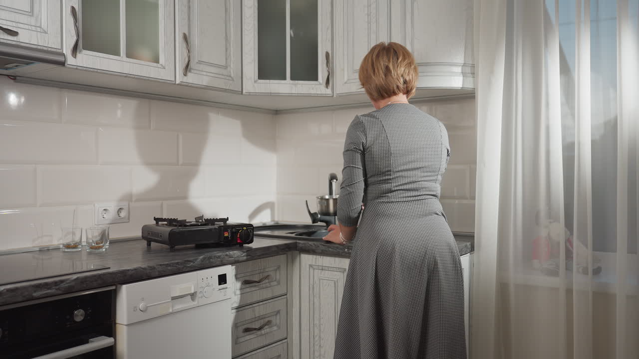 Woman in kitchen filling pot with water from tap, turning off tap once pot is full, captured from behind, with sunlight filtering through curtains and kitchen appliances around