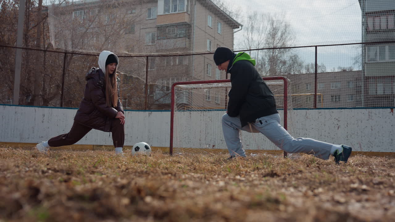 골대에서 공을 차지하기 위해 싸우는 백인 십 대, 거친 잔디밭에서 낮은 앵글, 수비수는 달려들고 공격수는 드리블, 투지 넘치는 야외 경기 분위기, 프레임 안에 아파트 단지와 네트, 높은 에너지와 투지
