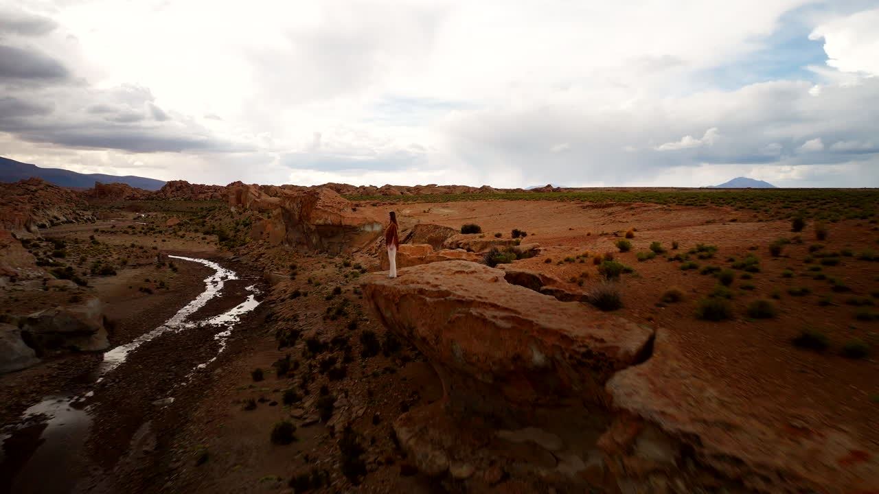 Woman and man stand overlooking dry river valley cutting through arid Sioli Desert landscape in Bolivia, dynamic aerial pullback