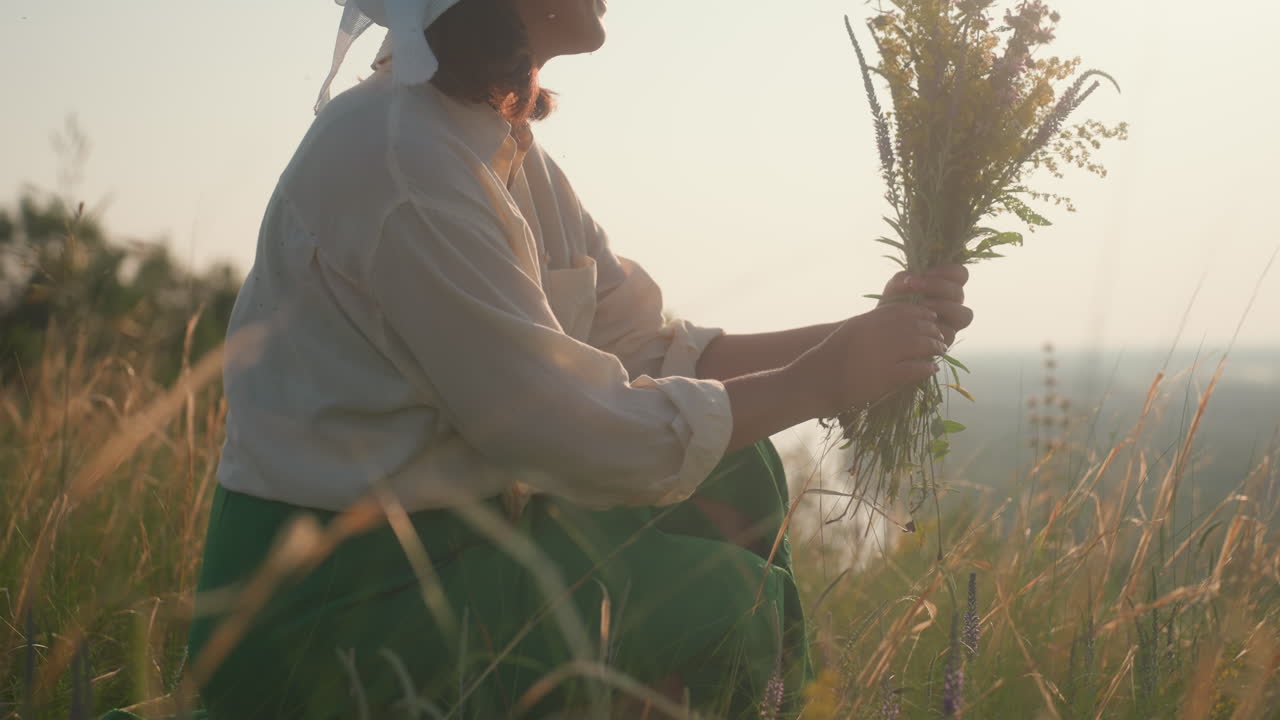 woman in green dress and white shirt kneeling in tall grass with relaxed smile while holding bouquet of wildflowers in warm sunlight, soft blurred background