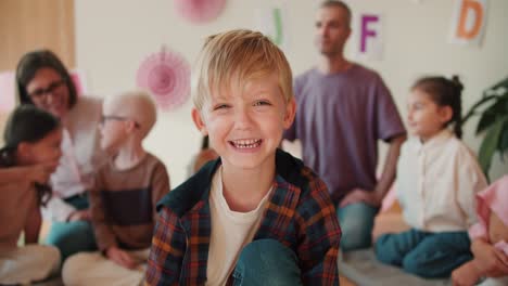 Porträt-Eines-Glücklichen-Und-Fröhlichen-Jungen-Mit-Blauen-Augen-In-Einem-Karierten-Hemd,-Der-Vor-Dem-Hintergrund-Seiner-Ersten-Unterrichtsstunde-Sitzt-Und-Lächelt,-Um-Sich-Zusammen-Mit-Seinen-Lehrern-Und-Einer-Gruppe-Von-Kindern-Auf-Die-Schule-Vorzubereiten