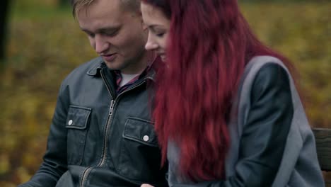 Cute-couple-sitting-on-a-bench-in-autumn-park-and-using-a-digital-tablet-together.-Young-woman-with-red-hair-and-attractive-man-in-a-leather-jacket-discussing-someting