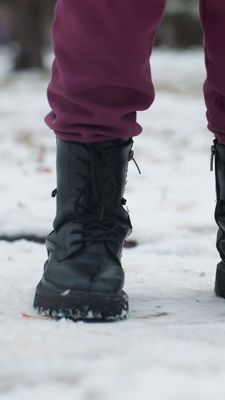 primer plano de piernas con botas negras mientras una persona salta con una pierna en el suelo cubierto de nieve, la nieve se desplaza ligeramente bajo los pies, creando un movimiento sutil