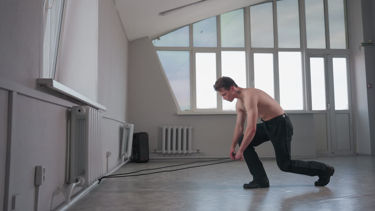 shirtless white man with neck tattoo strides across sunlit studio floor, picks up thick black rope lying coiled, secures rope around wrist and pulls with effort, emphasizing muscle tension