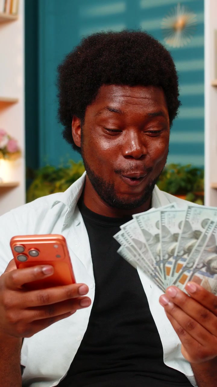 Giovane uomo afroamericano sul divano di casa che conta contanti con lo smartphone dopo una vincita a scommesse di giochi online.