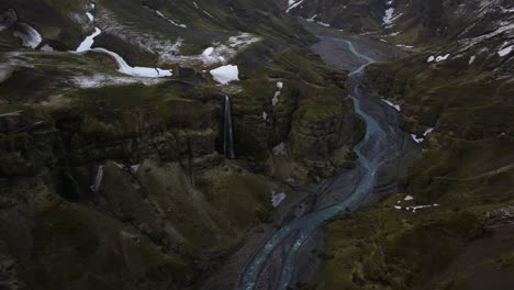 drone shot of a waterfall cascading into a river winding through búrfell canyon, iceland, surrounded by steep cliffs, snow patches, and rugged terrain