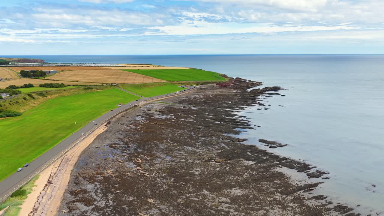 Drone glides above coastal road, green fields, rocky shoreline, and calm sea under daylight