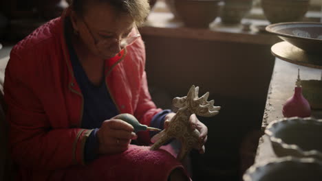 serious lady drawing on product in pottery. woman decorating clay toy in studio