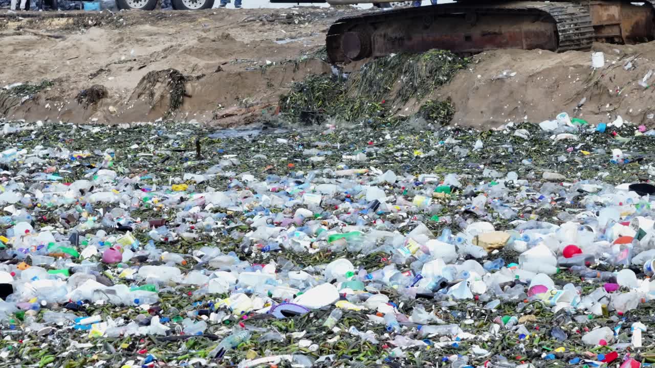 Waves near the shore, filled with plastic waste, contrast with an excavator tirelessly working to clean the area. An ideal scene for sustainability themes