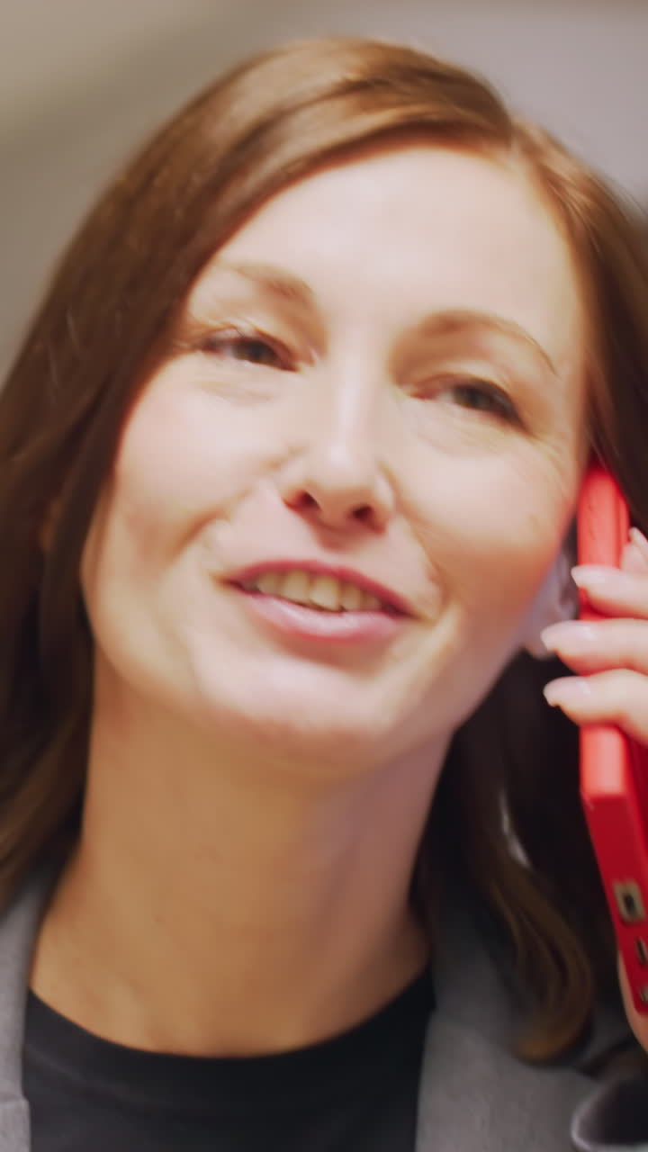 Businesswoman talking on phone in office corridor, looking focused and engaged. Holding red folder, wearing gray business suit. Modern office space with minimalist design in background
