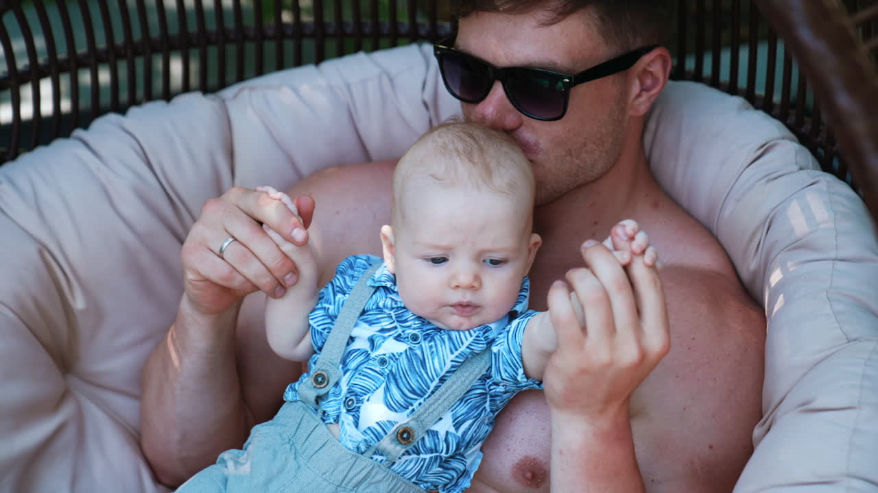 Little Caucasian newborn lying on dad's chest. Loving father kisses his son on the head sitting in egg chair outdoors.