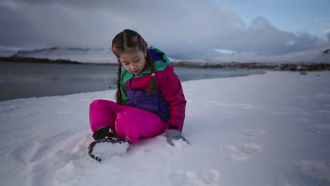 girl sitting on a snowy lakeside ground and enjoying peaceful winter mountain view while playing snowball