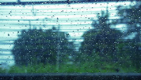 Raindrops-On-Window-With-Blurry-Trees-As-Background-During-Rainy-Day-In-Bangkok,-Thailand---close-up