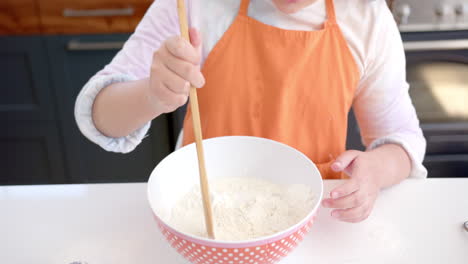 Happy biracial girl with long, curly hair mixing flour in bowl and smiling in sunny kitchen