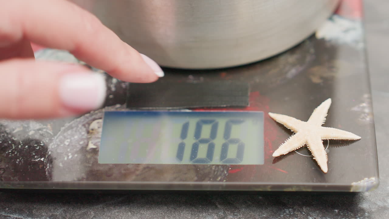 Close up of woman finger operating digital kitchen scale with container placed on top while display shows weight measurement, starfish decor sits on edge