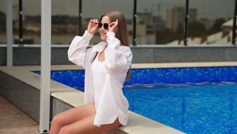 smiling happy girl wearing sunglasses enjoying sun at the swimming pool. lady in white swimsuit takes off glasses to sunbathe.
