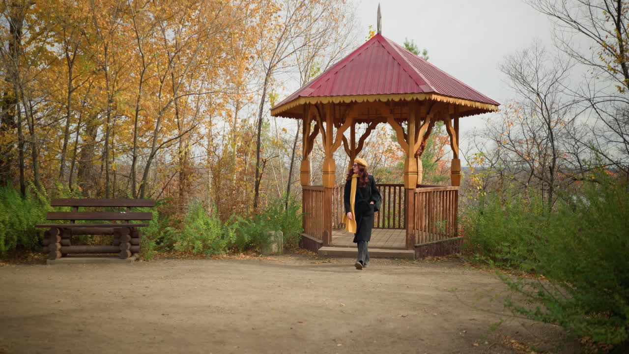 Lady in black coat, yellow scarf, and beret stepping out of wooden gazebo in serene autumn park, wooden bench nearby complements tranquil scenery of golden trees and fallen leaves