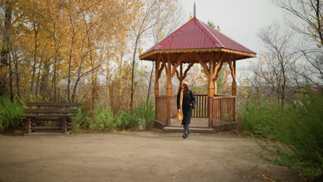 dama con abrigo negro, bufanda amarilla y boina saliendo de un mirador de madera en un sereno parque de otoño, un banco de madera cerca complementa el paisaje tranquilo de árboles dorados y hojas caídas