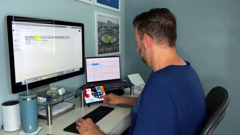 hombre blanco mayor chequeando ipad mini en el espacio de trabajo de la oficina en el hogar sentado frente a un gran monitor