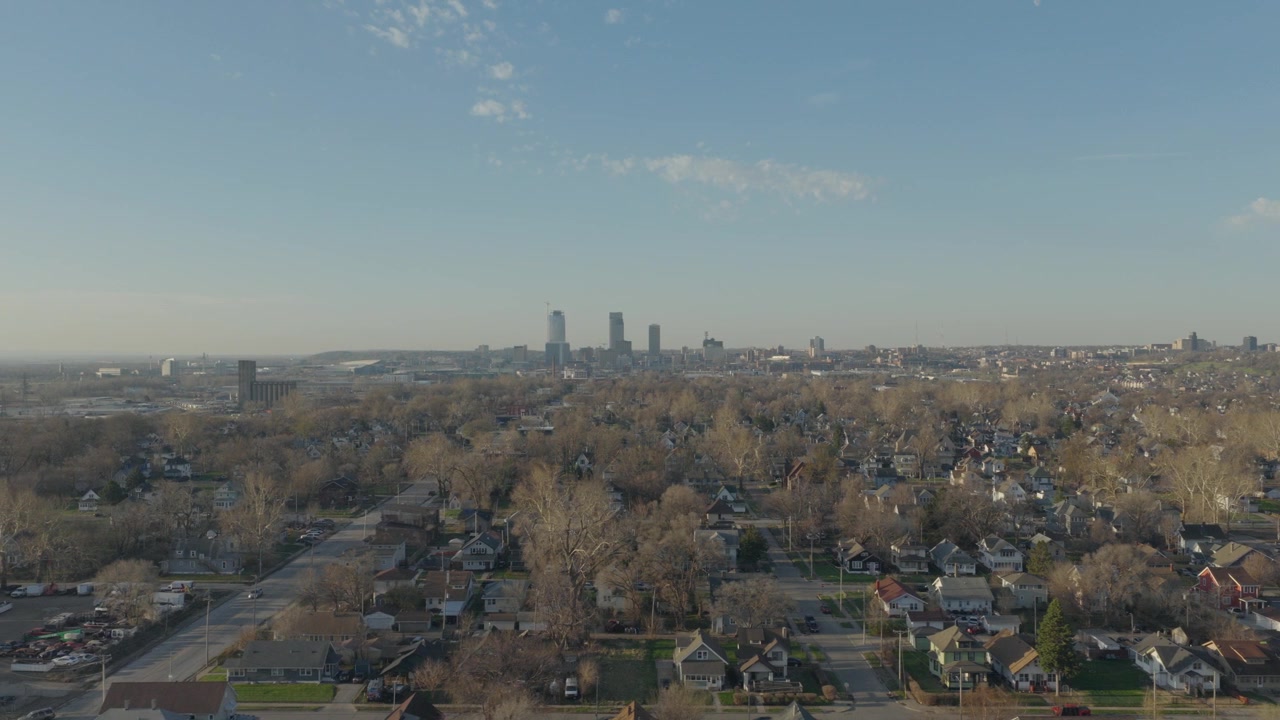 Sorvolo aereo laterale sopra i tetti del quartiere residenziale di North Omaha con alberi invernali spogli e lo skyline del centro di Omaha visibile all'ora d'oro.