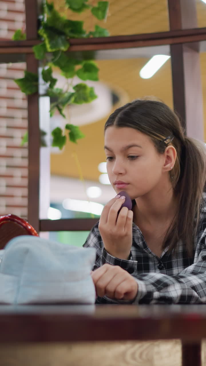 Adolescente aplicándose base de maquillaje con una esponja en una mesa de madera, camisa de cuadros y separador cubierto de plantas, expresión tranquila y concentrada, iluminación ambiental de centro comercial, espejo y polvos compactos cerca, movimientos constantes para difuminar.