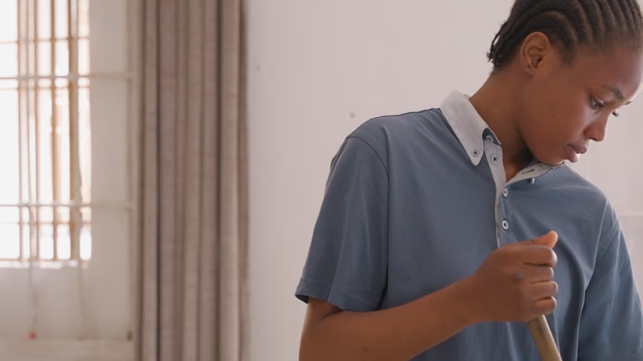 Adolescente nero che spazza il pavimento all'interno della casa, movimento a figura intera in una stanza soleggiata con finestra e tenda, polo casual, espressione determinata e concentrata, compito domestico ritmico, pulizia quotidiana