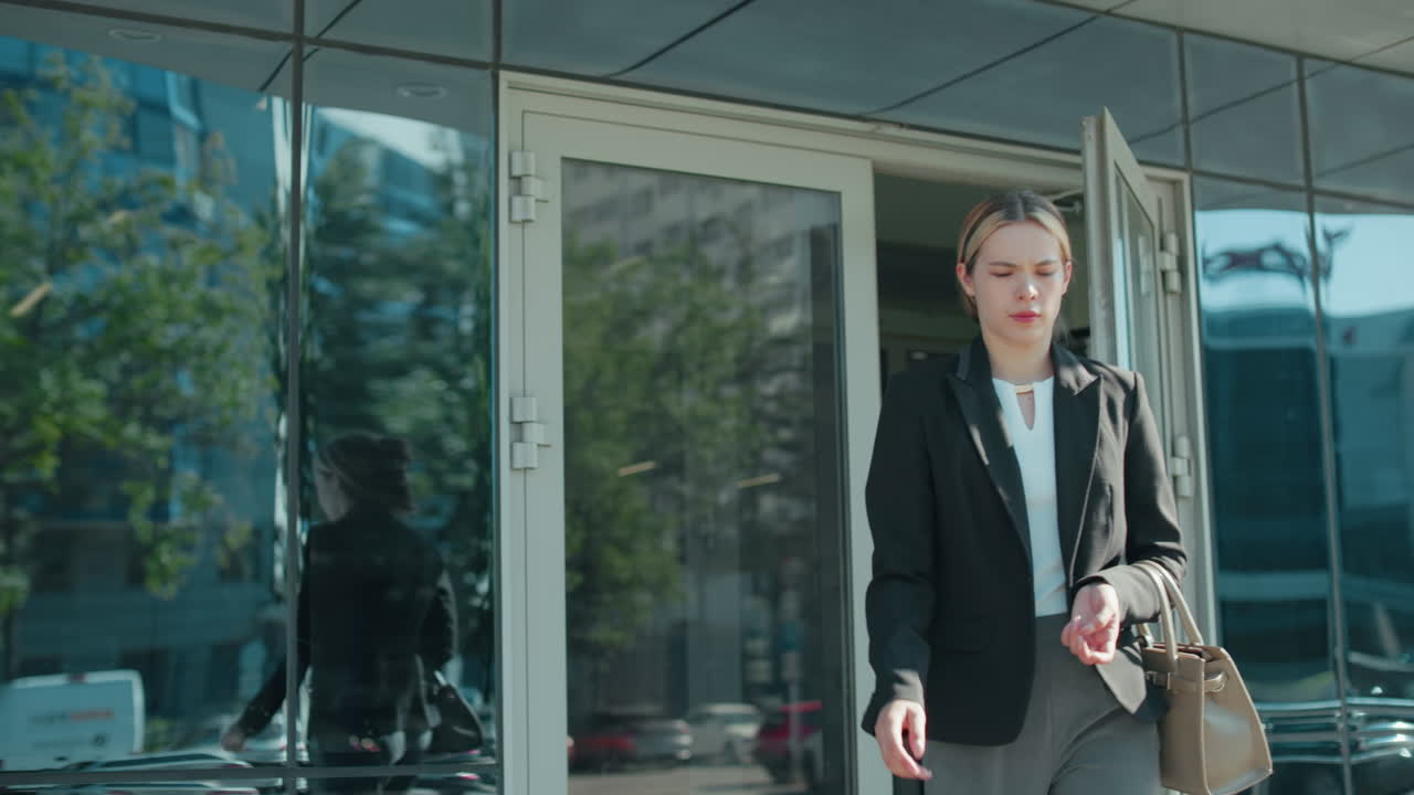 Business lady carrying handbag walks into office building dressed professionally in black jacket and gray pants, with glass door reflecting urban street, trees and parked cars