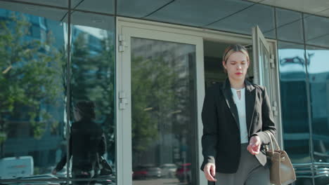 business lady carrying handbag walks into office building dressed professionally in black jacket and gray pants, with glass door reflecting urban street, trees and parked cars