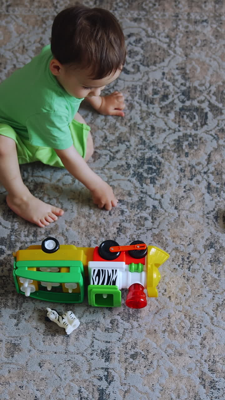 One year old toddler sitting on the carpet in the room. Dark-haired boy plays with the toy train. Top view. Vertical video