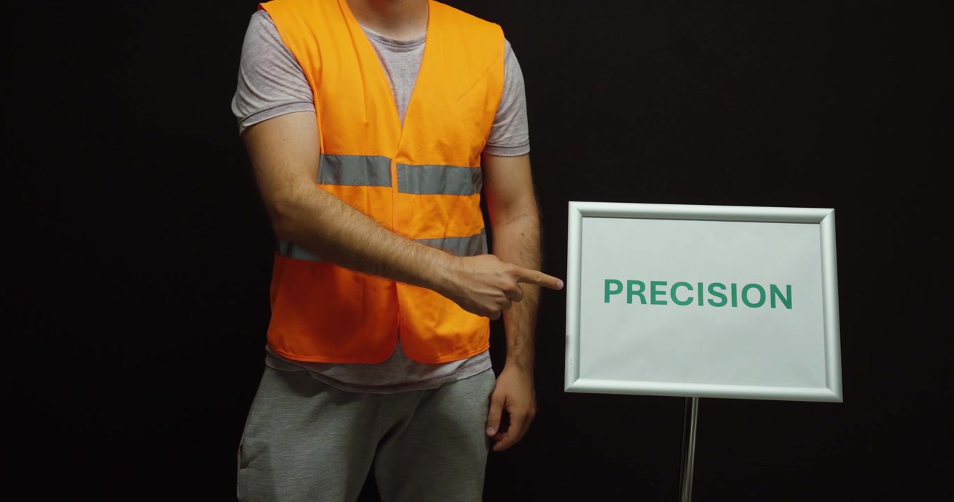 Trabajador con chaleco de seguridad señalando un letrero de precisión, que representa trabajo preciso, calidad, fabricación, ingeniería y atención al detalle.