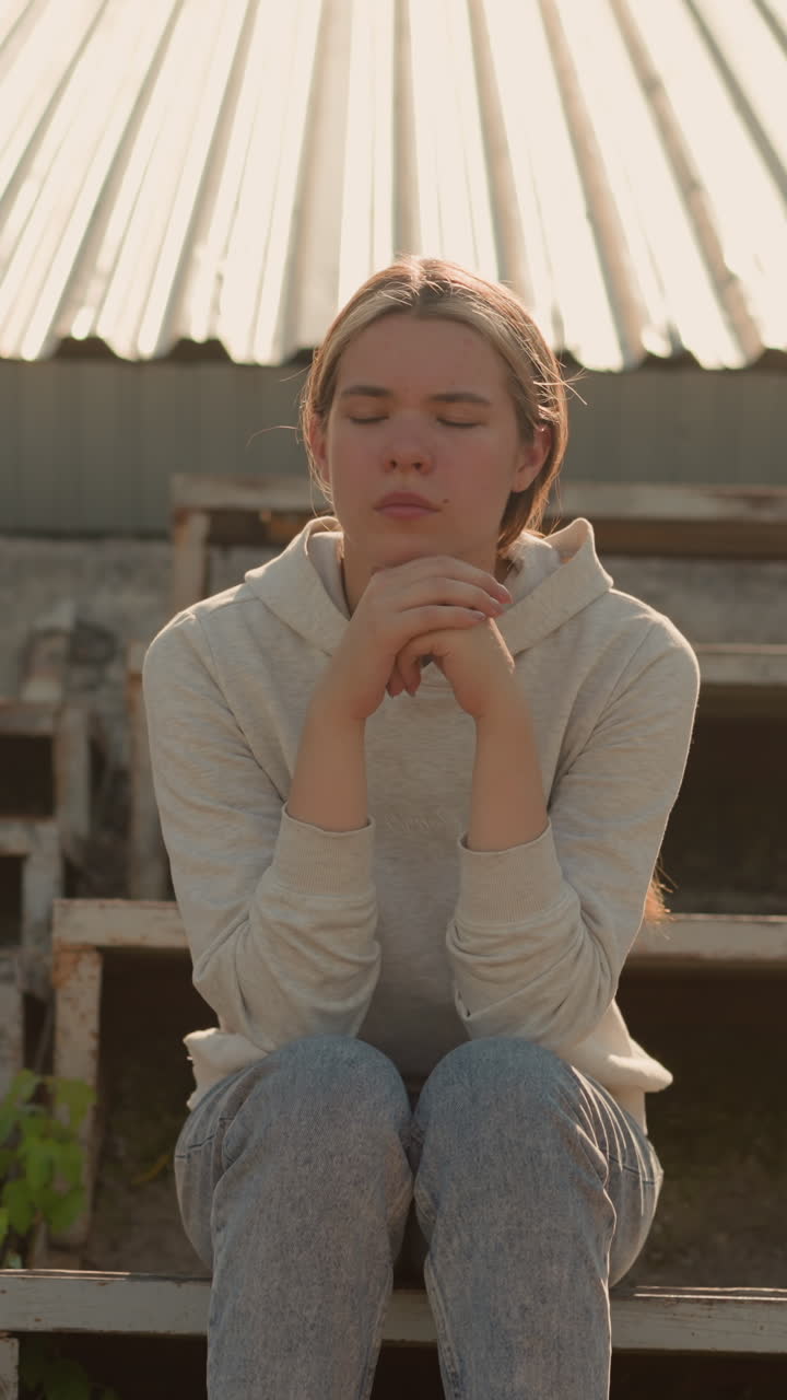 Thoughtful woman in casual attire sits on rustic stadium bleachers, hands joined together, with a pensive expression, looking contemplative in the soft outdoor light