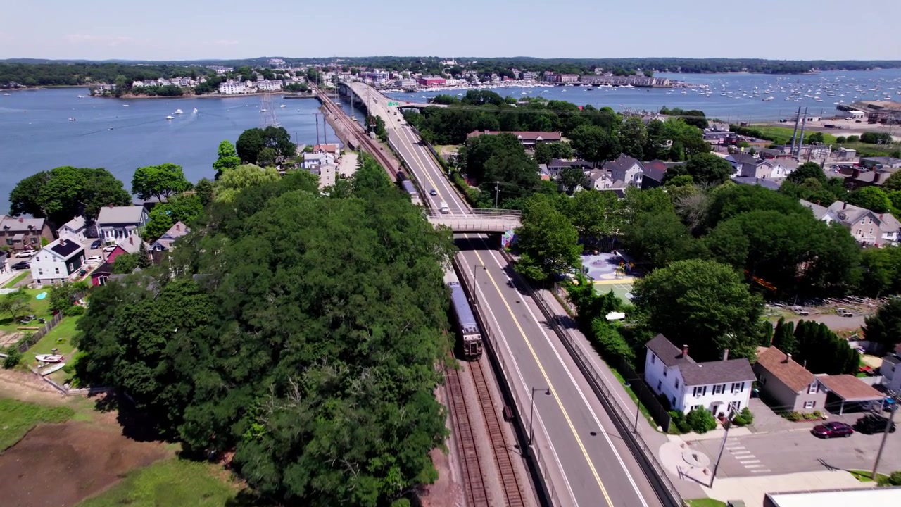 Un treno pendolare viaggia lungo un fiume e una strada trafficata a Salem, Massachusetts. Un ponte e un grande serbatoio di carburante commerciale sono visibili in lontananza