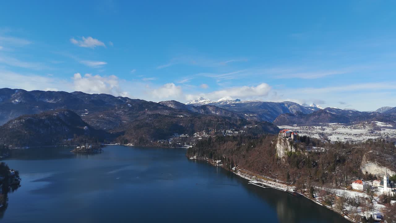 Panorama aereo invernale del Lago di Bled, Slovenia, con il Castello di Bled sopra l'acqua blu, la riva innevata e le montagne alpine sotto un cielo limpido e luminoso.