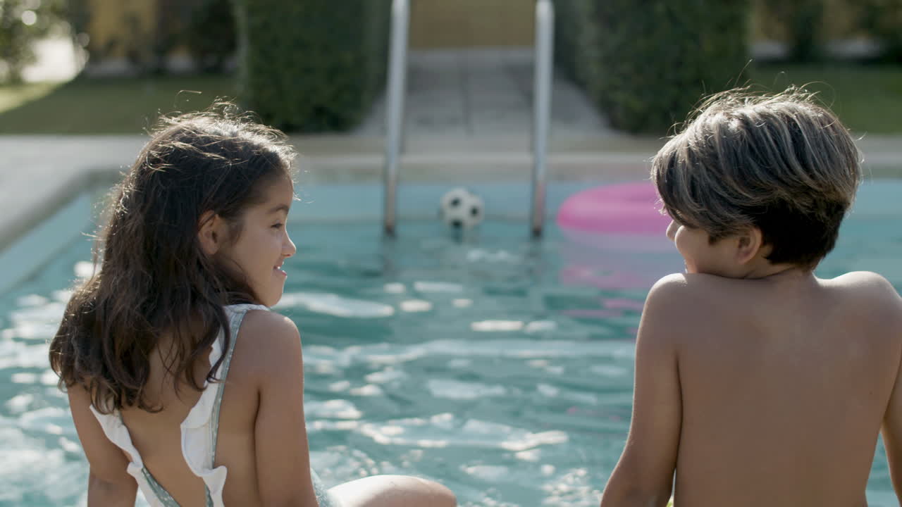 vista de atrás de un niño y una niña sentados en el borde de la piscina en un día soleado.