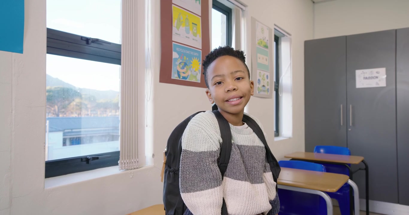 Ragazzo preadolescente che risponde alla telecamera, sorridendo e parlando in classe con uno zaino, indossando un maglione.