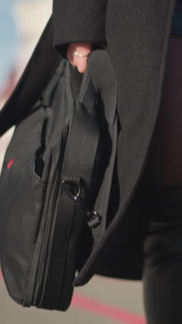 primer plano de una mujer elegante caminando con confianza por una calle urbana iluminada por el sol, con abrigo negro y botas de cuero, llevando dos bolsas, con la pierna revelada debajo del abrigo, el fondo presenta un edificio borroso