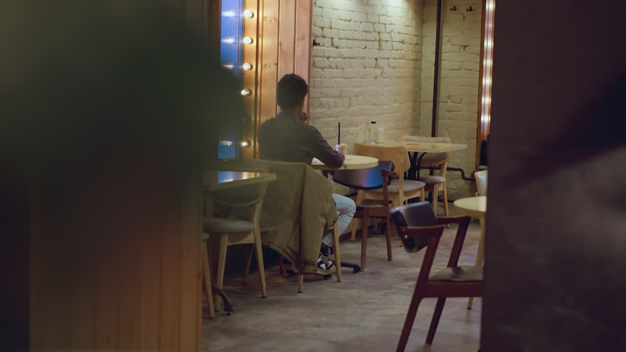 Man in gray shirt sitting alone with drink in hand near window with mirror lights in empty cozy caf , facing wall with white bricks, soft ambient glow creating quiet urban evening atmosphere around him