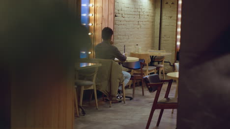 man in gray shirt sitting alone with drink in hand near window with mirror lights in empty cozy caf , facing wall with white bricks, soft ambient glow creating quiet urban evening atmosphere around him