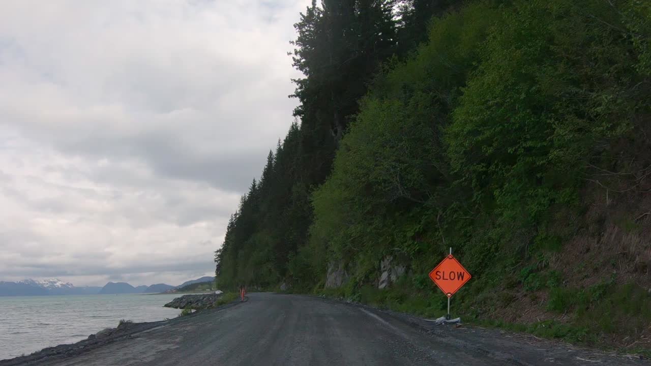 POV - Driving on Lowell Point Road that is a gravel road that runs along a tree-covered mountain side and drops off to the ocean bay; near Seward, Alaska