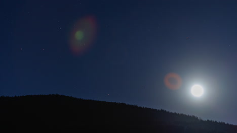 Glowing-bright-moon-rises-over-wooded-mountains-with-deep-blue-sky-and-stars