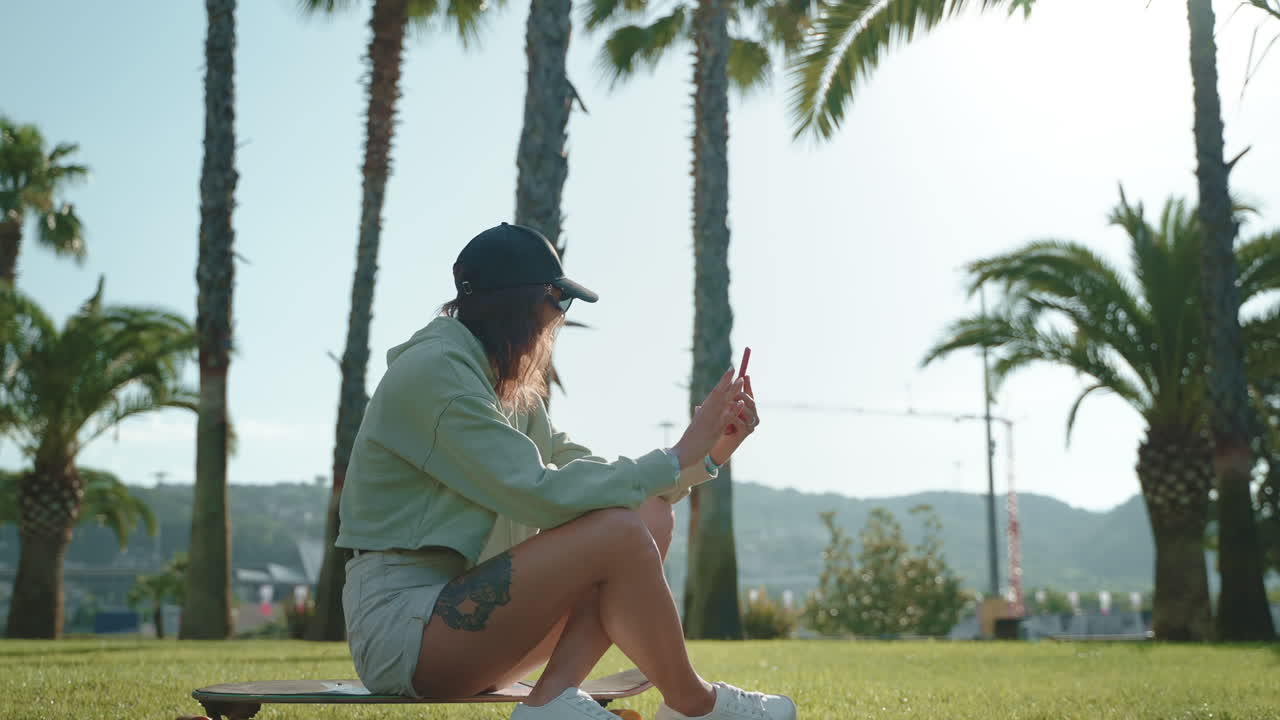 스케이트보드 에 앉아 있는 여자 가 공원 에서 전화 를 사용 하고 있다
