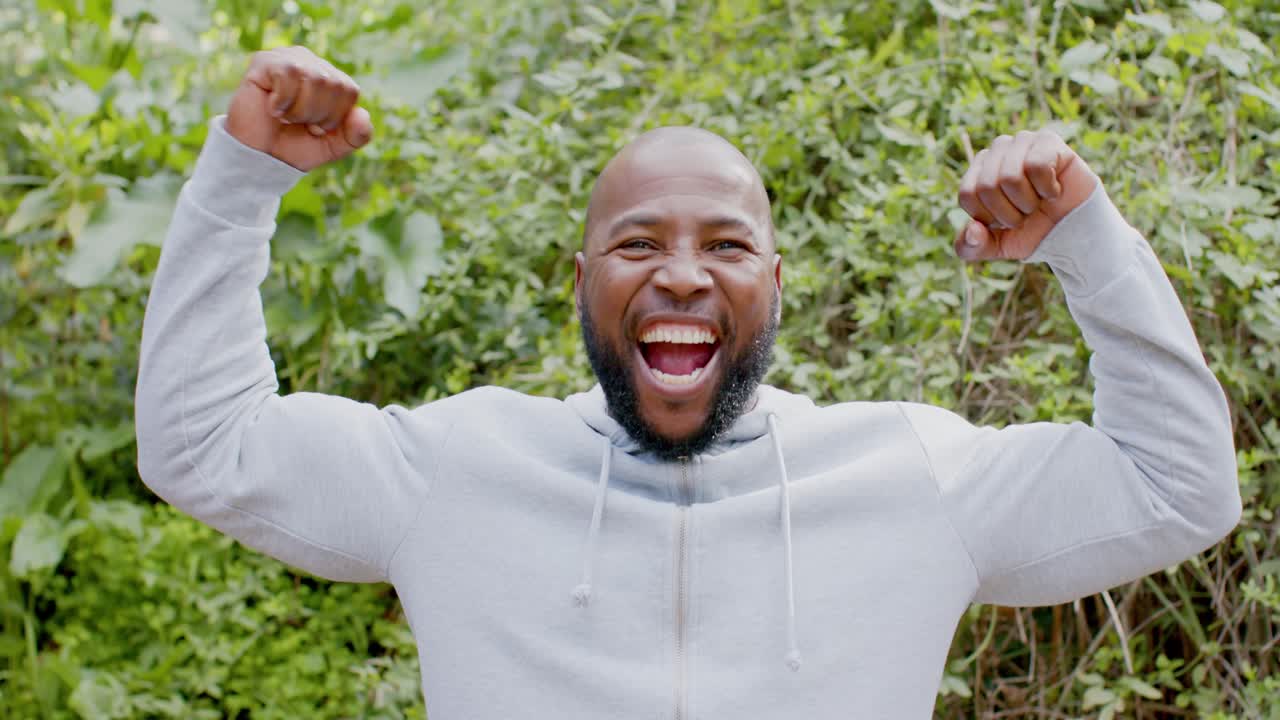 Mature African American man greeting viewer jogging in place outdoors, flexing biceps and cheering