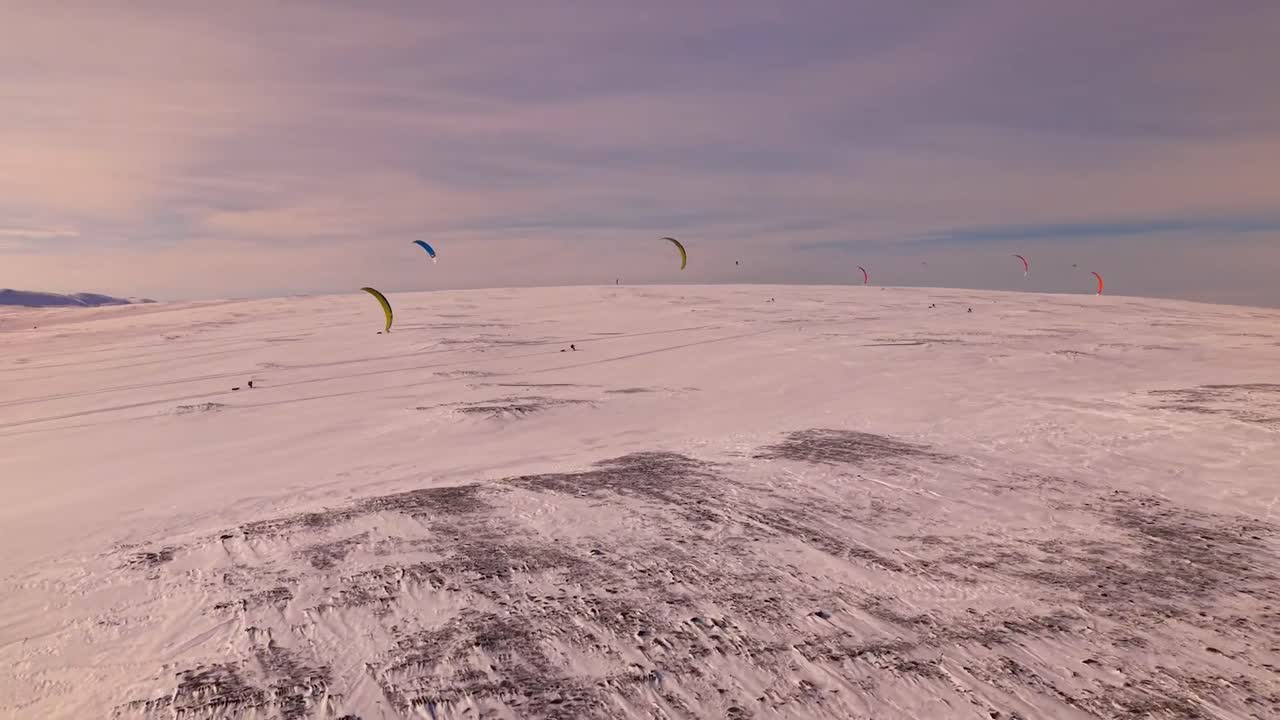 Aerial drone footage panning slowly and moving forward over white snow covered Norway landscape where people are doing an extreme sport endurance race with kiteskiing or snowboarding with kites, sunny