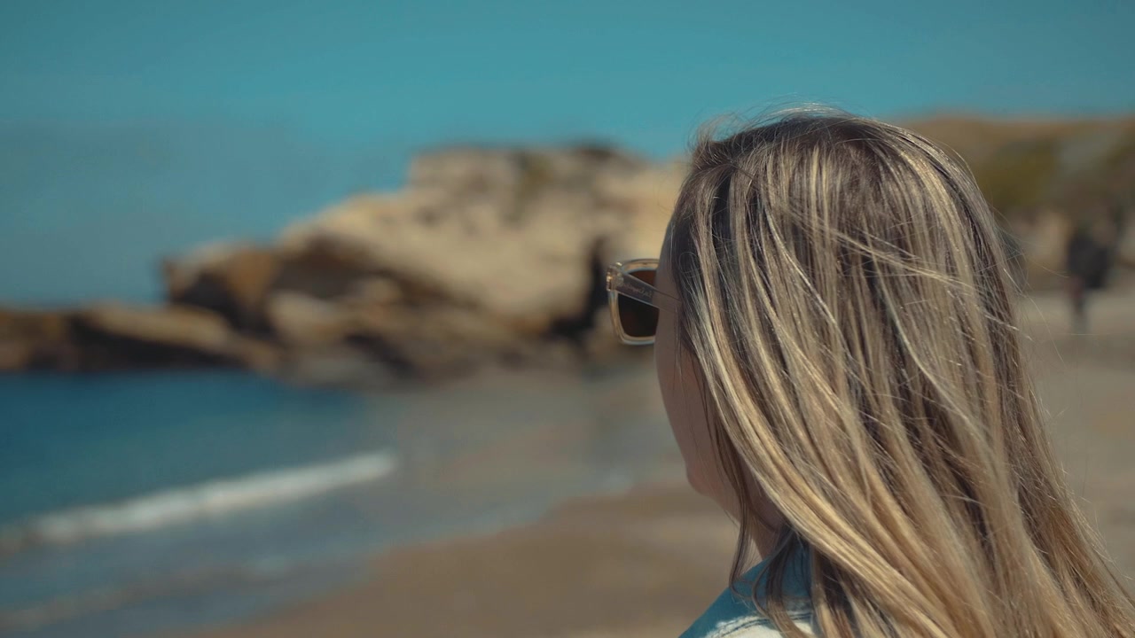 Blonde Woman in Sunglasses Looking at the Ocean from a Sandy Beach